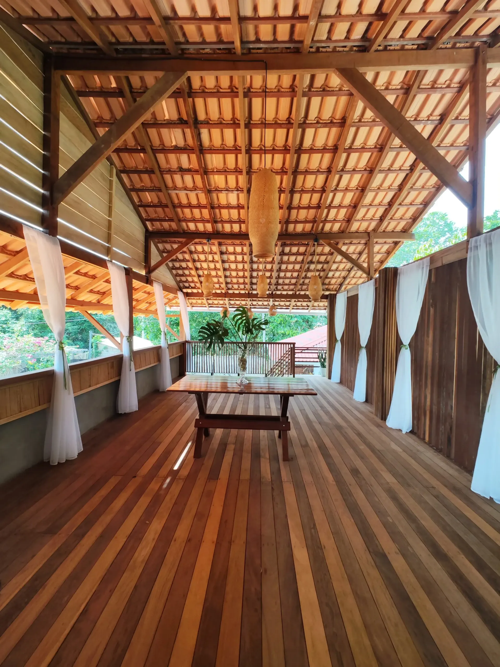 Deck de madeira no andar superior, com estrutura de telhado aparente e cortinas brancas abertas para as árvores